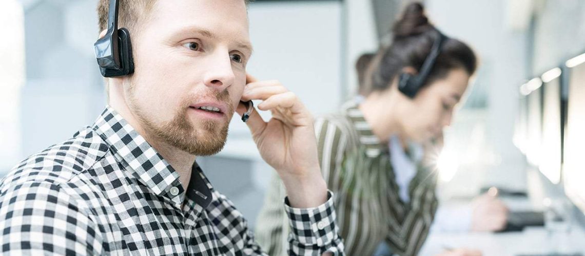 Portrait of  young man wearing headset working with group of help desk operators sitting in row and  talking to clients , copy space